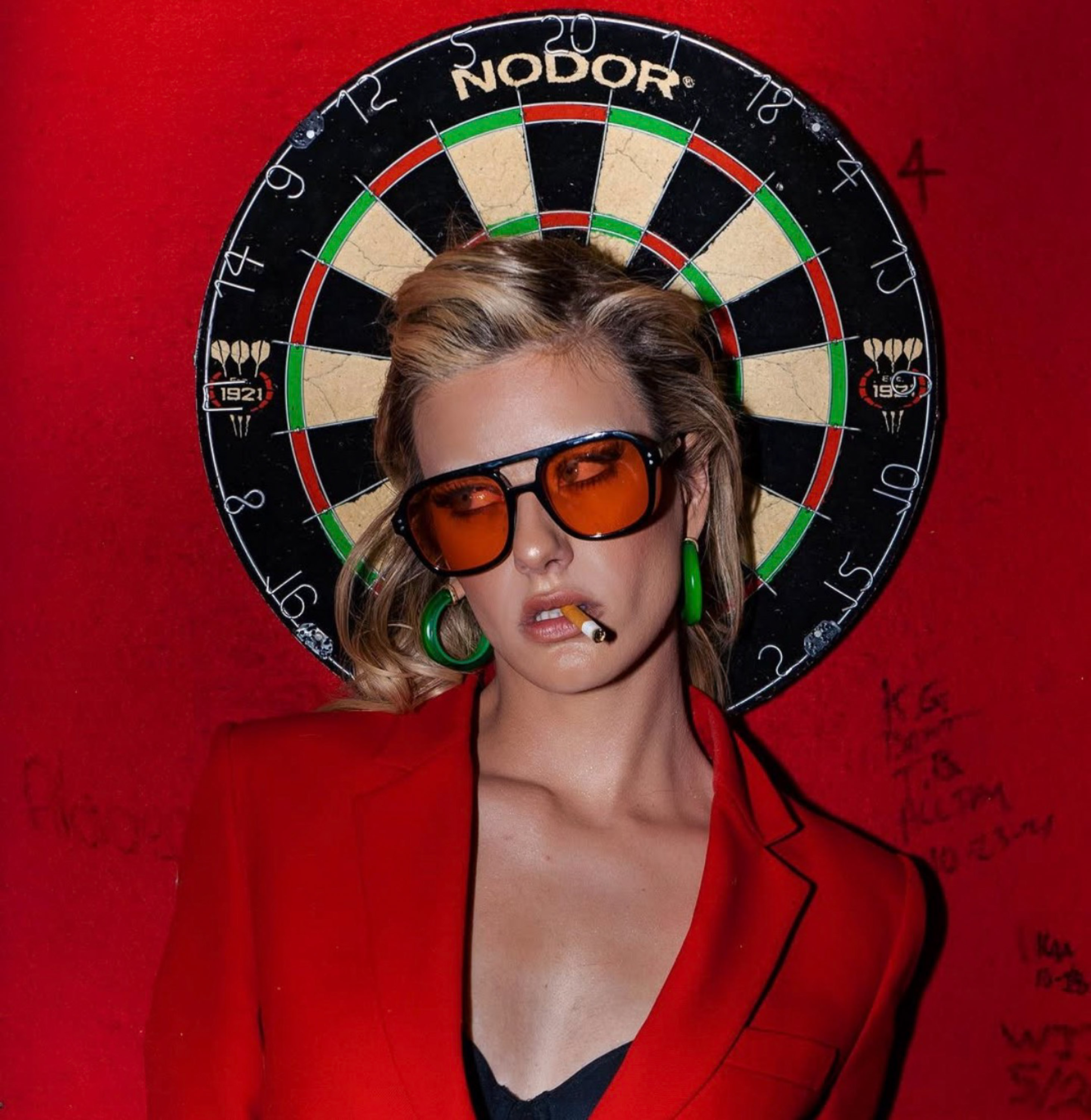 Dawn Muensterman in a red blazer and amber sunglasses in front of a dartboard on a red wall